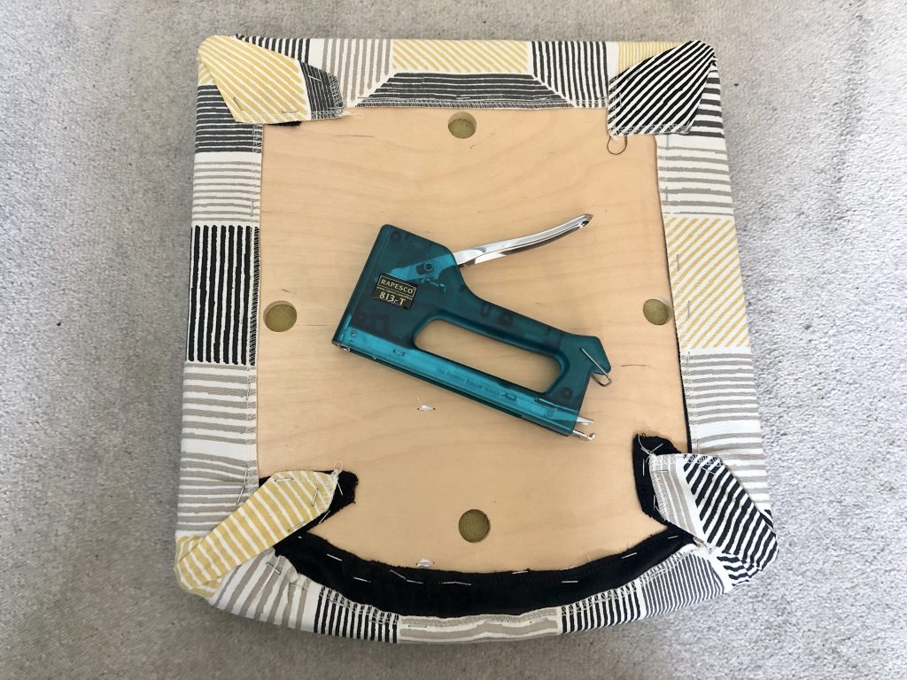 Chair pad covered in fabric and stapled in place, a green staple gun sits in the middle of the photo