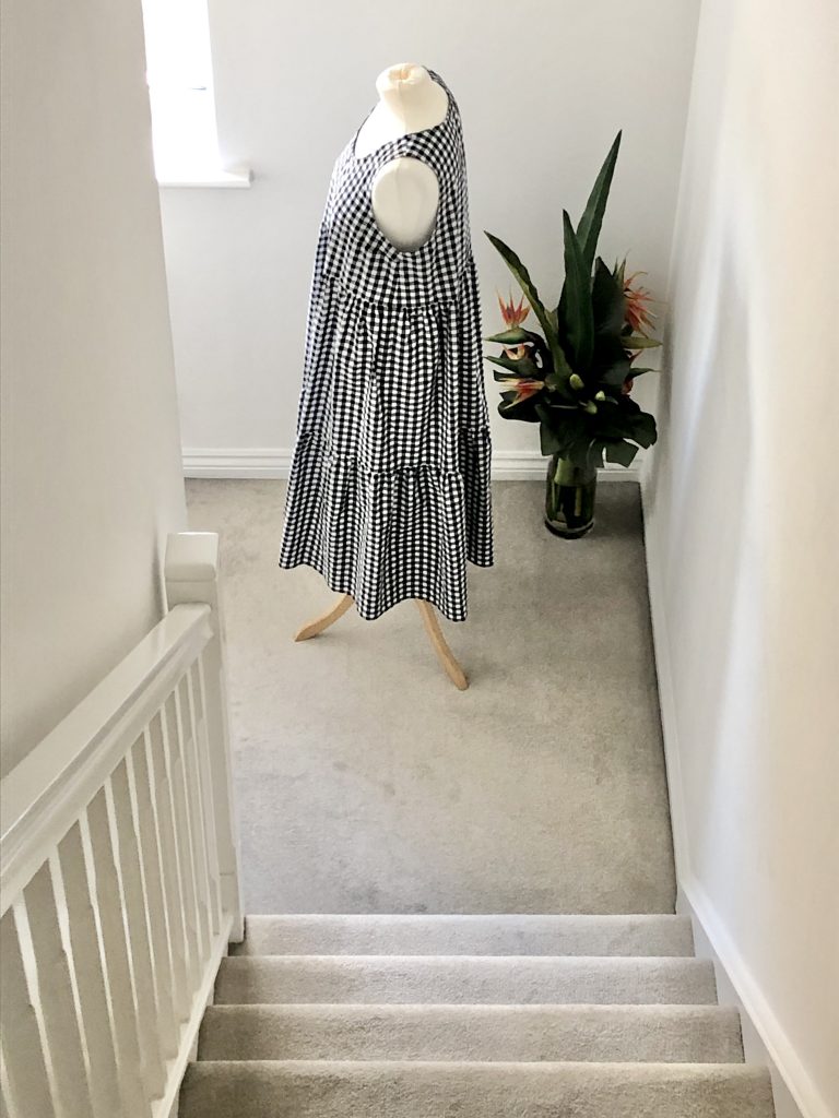 View down a carpeted staircase to the Verano dress - a black and white gingham dress on a white mannequin next to a house plant