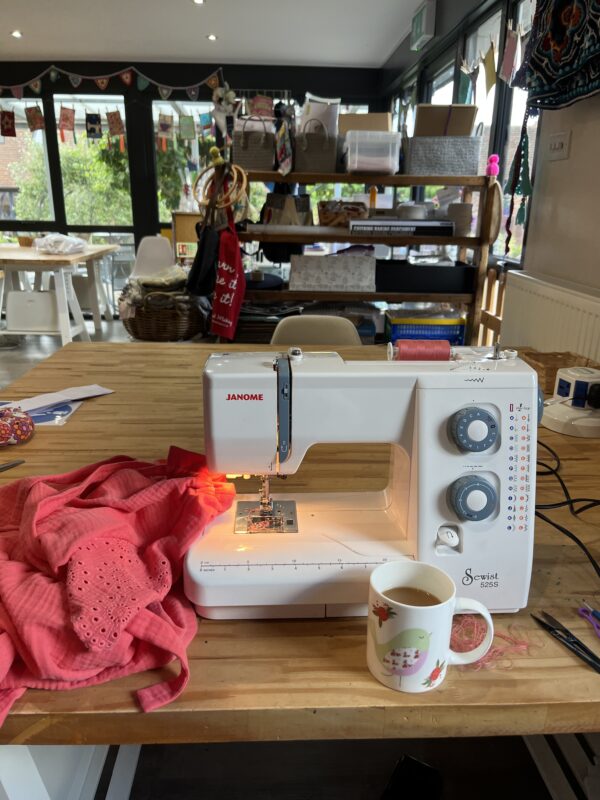 a sewing machine, mug of tea and coral coloured fabric are on a wooden table in the made and making studio