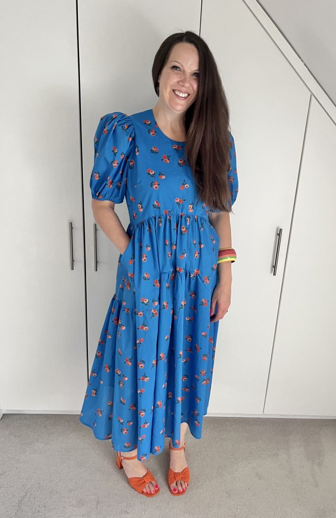 Julia, a tall woman with long brown hair, is wearing a mid length blue dress with orange flowers on it. She is wearing orange shoes and a striped bracelet.