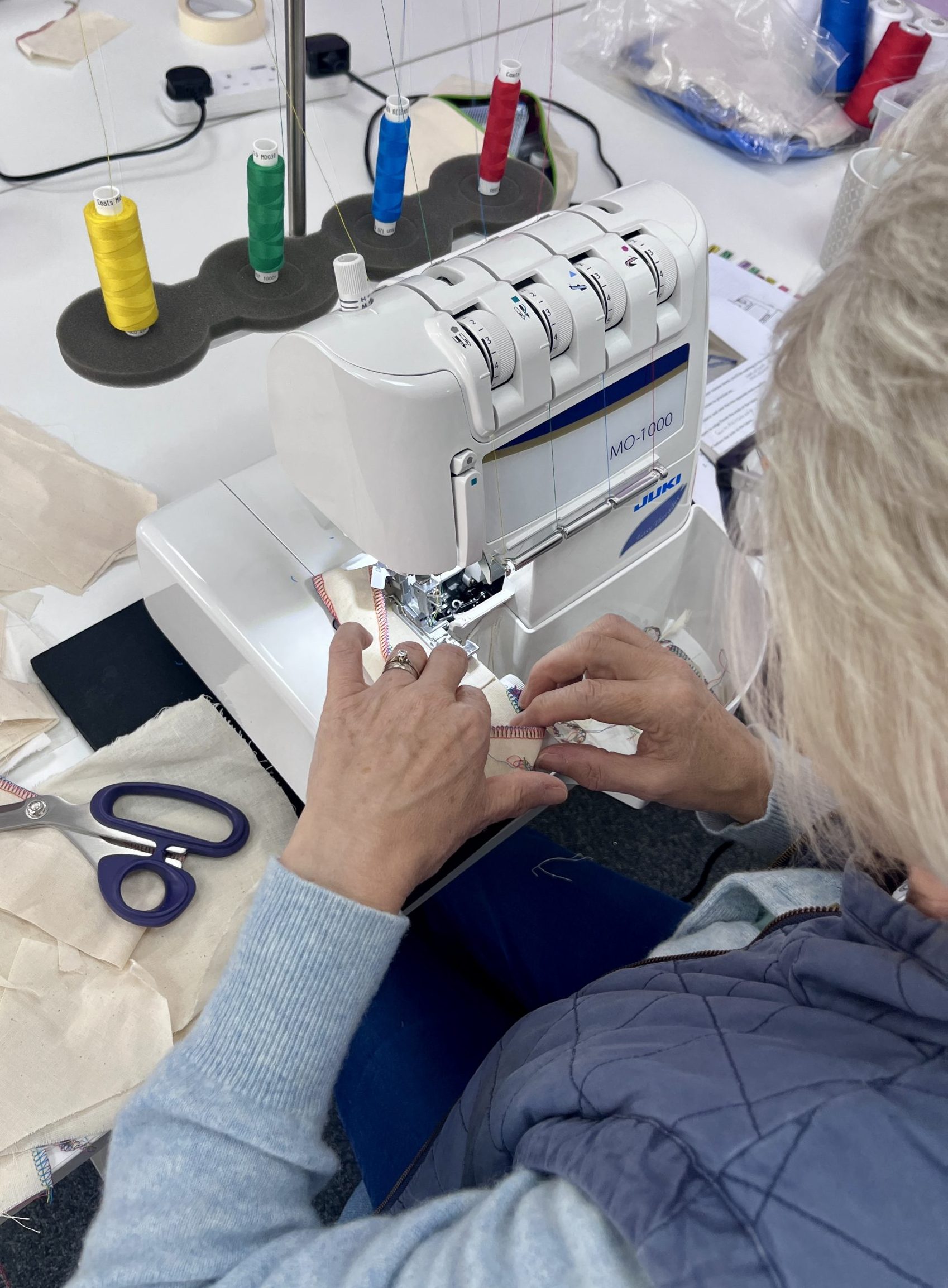 A woman with blond hair is operating an overlocker sewing machine. Spools of yellow, green, blue and red sit on top of the machine.