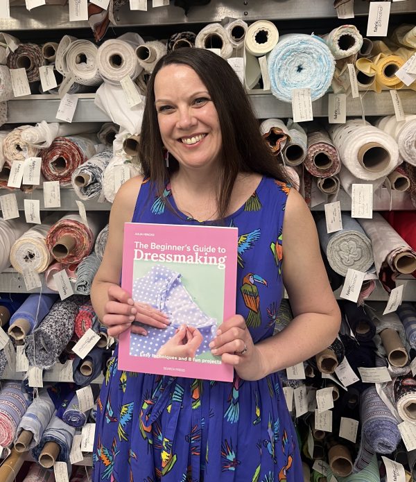 Julia is wearing a sleeveless dress with neon parrots on it and is holding her book 'The Beginner's Guide to Dressmaking'