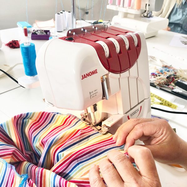 an overlocker is threaded with blue fabric. a pair of hands are feeding striped fabric through the machine