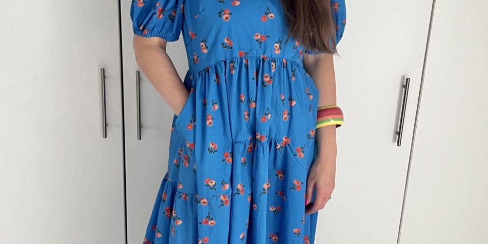 Julia, a tall woman with long brown hair, is wearing a mid length blue dress with orange flowers on it. She is wearing orange shoes and a striped bracelet.